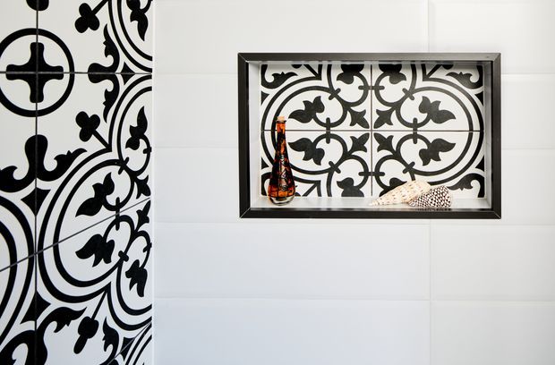Close-up of a refined bathroom wall featuring large white wall tiles and a bold black-and-white ornamental patterned tile installation along the left side. A recessed, dark-framed display niche contains a decorative bottle and shell, with the same patterned tile used as its backing.