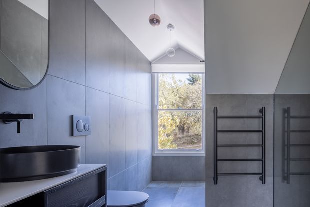 A compact contemporary bathroom with a dark grey tiled or panelled wall finish, black vessel basin on a light benchtop, wall-mounted black tapware, integrated toilet, mirrored cabinet, and black heated towel rails. A high sloping ceiling with pendant lights and a tall window bring natural light into the space.
