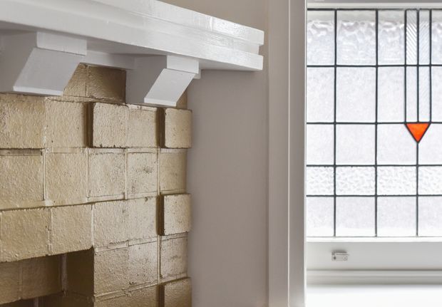 Close-up architectural detail of a traditional masonry fireplace with cream painted or rendered brickwork, a white timber mantel with chunky brackets, and an adjacent leadlight window featuring geometric panes and a small orange-red diamond accent. The image emphasizes heritage-inspired fixed architectural elements rather than a complete room.
