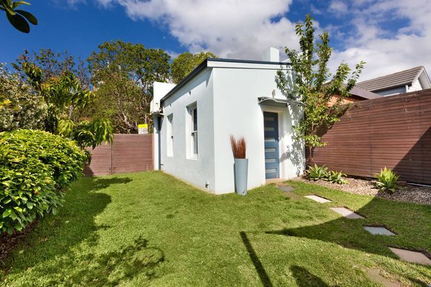 A small detached white rendered building with a flat roof and narrow vertical windows, set within a landscaped suburban garden. A partially glazed entry door sits beneath a small curved awning, with stepping-stone paths, lawn, timber privacy fencing, and established planting surrounding the structure. A neighbouring pitched-roof building is visible beyond the fence.