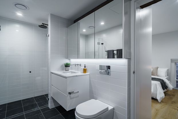 A bright, contemporary bathroom with white rectangular wall tiles, dark tiled flooring, a frameless glass shower, wall-mounted white vanity, toilet, large mirror, and recessed ceiling lights. A bedroom is visible through the open doorway on the right, with timber flooring and a partially visible bed.