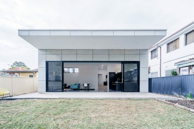 Front-on exterior view of a minimalist single-storey extension or pavilion with a broad flat roof, pale large-format facade panels, deep roof overhang, and expansive dark-framed sliding glass doors. A sparse contemporary living area is visible inside, opening onto a lawn; neighbouring suburban homes and fences frame the property.