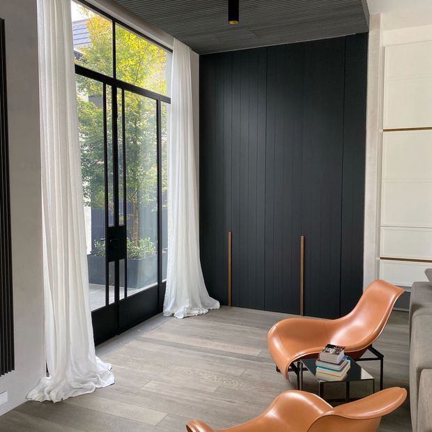 A refined contemporary living area with floor-to-ceiling black-framed glazing and sheer white curtains opening to a small balcony or terrace. A dark vertical-panel feature wall with tall integrated cabinetry contrasts with pale timber-look flooring and white built-in joinery. Two sculptural tan leather lounge chairs and a small black side table create a sophisticated seating nook. The ceiling has dark linear panelling and recessed lighting.