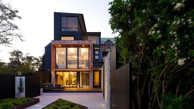 Contemporary multi-storey residential home viewed from the front, with dark cladding, angular roof forms, extensive glazing, warm interior lighting, and a paved entry path bordered by tall privacy fencing and established planting.