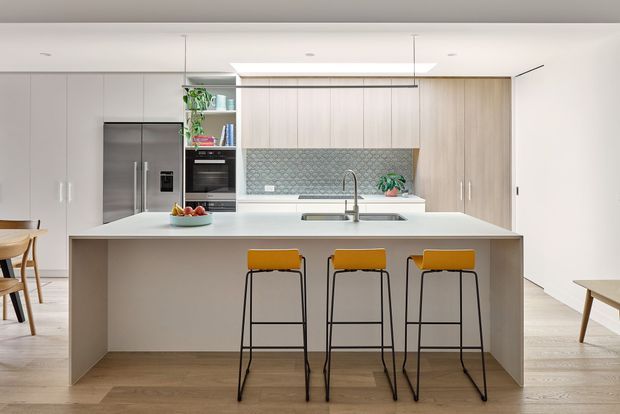 Bright contemporary kitchen with a large white island and three bar stools, pale timber cabinetry, integrated stainless-steel appliances, and a green patterned tile splashback. The open-plan space has light timber flooring, white walls, recessed ceiling lighting, and minimalist built-in joinery.