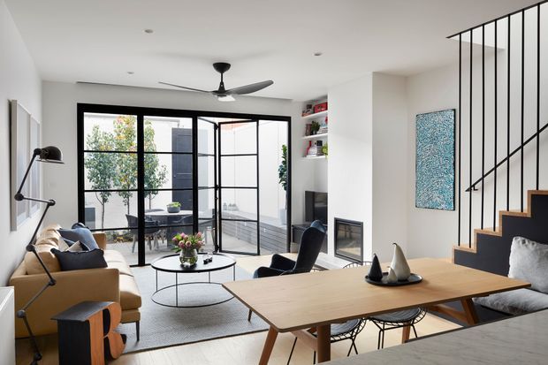 Bright open-plan living and dining area with a contemporary minimalist interior. The room features a large timber dining table with modern wire-frame chairs, a tan sofa, round coffee table, dark lounge chairs, built-in shelving, a linear fireplace, and black-framed glazed doors opening to an outdoor deck. A black metal stair balustrade and timber stair treads rise along the right side. The palette is predominantly white, light timber, tan, black, and grey, with abundant daylight and a strong indoor-outdoor connection.