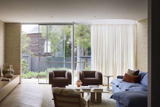 A bright, refined living room with pale exposed brick walls, full-height glazed doors and windows opening to a leafy courtyard, and sheer curtains. The room contains a blue sofa, two dark upholstered armchairs, a low timber and glass coffee table, a sculptural lounge chair, and understated decorative objects. The flooring is warm-toned timber laid in a chevron or herringbone pattern, creating a calm contemporary interior with strong indoor-outdoor connection.