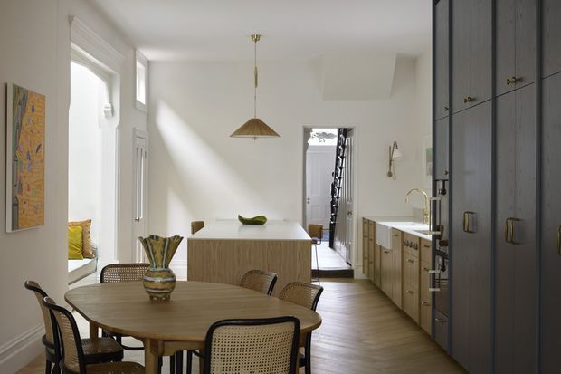 Bright open-plan kitchen and dining area with a round timber dining table and woven cane chairs in the foreground. A large light-wood kitchen island, integrated cabinetry, sink, and long run of cabinetry occupy the right side, while tall windows and a glazed door bring in strong natural light. Dark floor-to-ceiling cabinetry provides contrast, and a simple pendant hangs above the island.