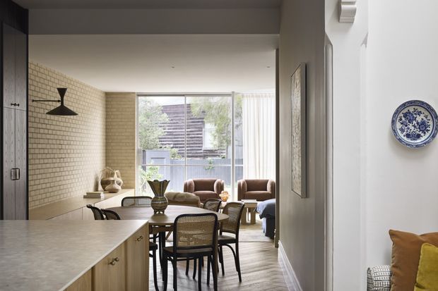 View from a kitchen through a dining area into a compact living room. The space features pale stone kitchen surfaces and light timber cabinetry, exposed cream brick walls, timber flooring, a simple dark pendant, a round dining table with woven-back chairs, and large glazing at the rear. The interior has a restrained contemporary design with warm, traditional decorative touches.