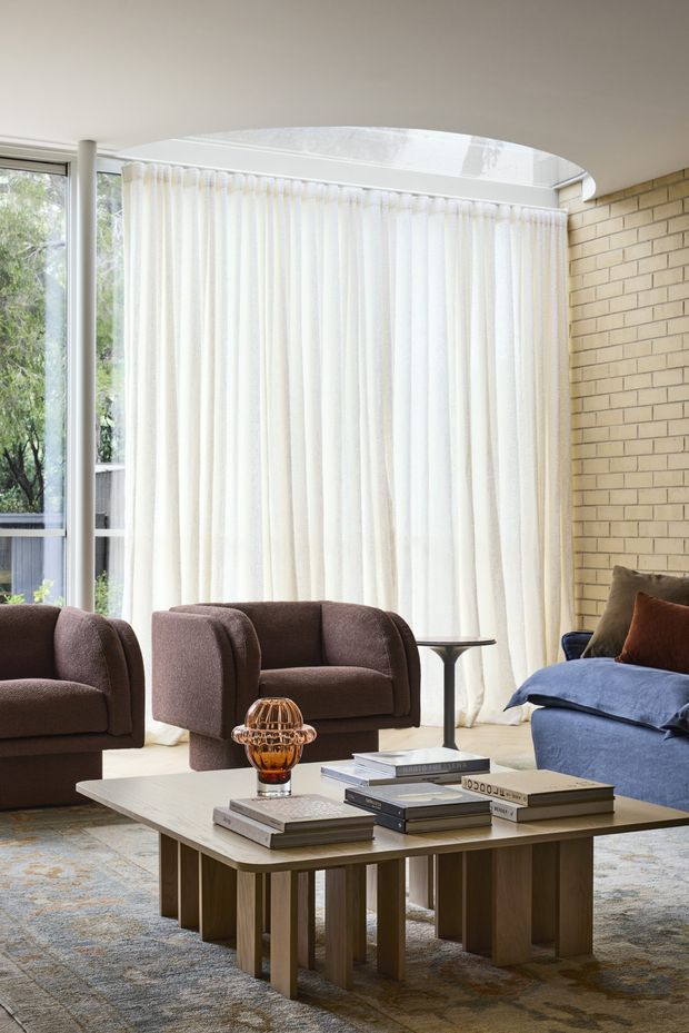 A bright, refined living room with a blue sofa, two brown upholstered armchairs, a sculptural wood coffee table stacked with books, and a small amber glass decorative lamp. Floor-to-ceiling sheer curtains cover broad glazing, while a curved skylight or roof opening brings in additional daylight. A warm pale brick feature wall contrasts with the neutral carpet and white ceiling.