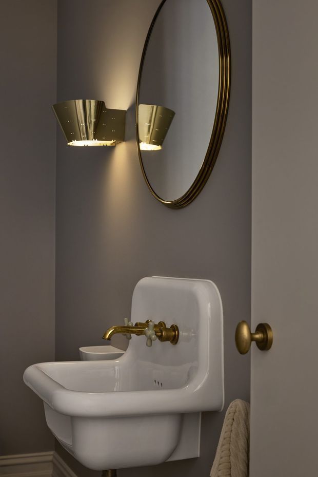A close-up of a compact, elegantly styled bathroom featuring a white wall-mounted basin with rounded porcelain forms, polished brass tapware and controls, a matching brass towel hook, an oval mirror with a layered metallic frame, and a sculptural brass wall sconce. The walls are painted a warm taupe-grey, with traditional decorative skirting visible at the base. The lighting is warm and atmospheric, creating a refined, luxurious mood.