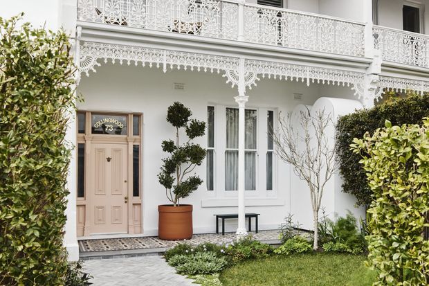 Front facade of a two-storey white residence with an ornate cast-iron balcony and decorative verandah, pale paneled entry door, tall windows, manicured hedges, topiary, and a patterned paved path. The architecture has a strongly Victorian character with intricate lacework detailing.