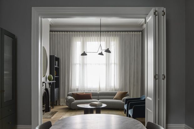 View through a wide open doorway into an elegant living room, with a curved grey sofa, sculptural pendant lights, full-height sheer and draped window coverings, dark built-in shelving, and a round dining table in the foreground. The room has refined traditional architectural detailing, including layered crown moulding, substantial door trim, and tall skirting boards, combined with contemporary furnishings in a restrained grey, cream, and dark palette.