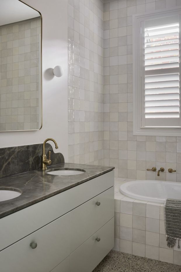 A bright, traditional bathroom featuring a tiled corner bathtub, double vanity with pale cabinetry, dark veined stone benchtop, brass tapware, a rounded-edge mirror, and a white louvred window. The walls and bathtub surround are clad in small glossy square tiles, while the floor has a speckled terrazzo-like finish.
