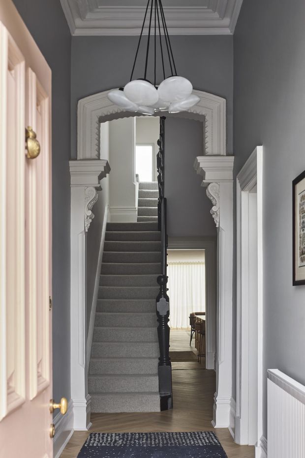 A formal residential hallway focused on an ornate staircase. The narrow corridor has dark grey walls, white decorative plaster mouldings and a coffered ceiling, with a carpeted stair rising through an elaborately trimmed archway. A black sculptural pendant light hangs overhead, while a dining area is visible beyond the stair and framed artwork and a radiator line the hall.