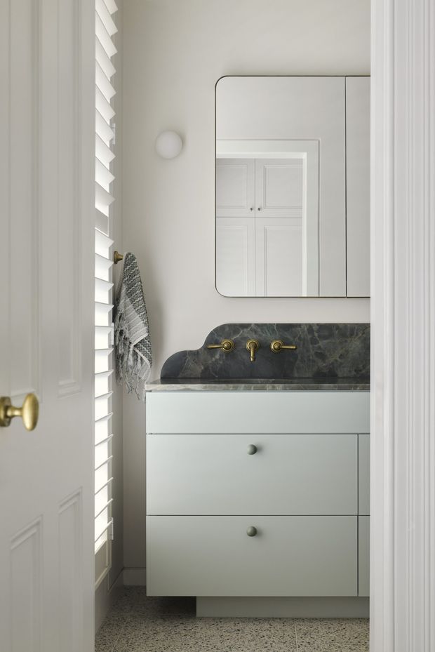 A compact, refined bathroom viewed through a partially open paneled door. The room features a wall-mounted pale vanity with integrated drawers, a dark veined stone backsplash and benchtop with sculpted raised side profile, brass wall-mounted tapware, a rounded-rectangle mirrored cabinet, white paneled doors, a wall sconce, louvred window shutters, and a terrazzo-style floor. The palette is predominantly soft white, pale grey, dark stone, and brass accents.