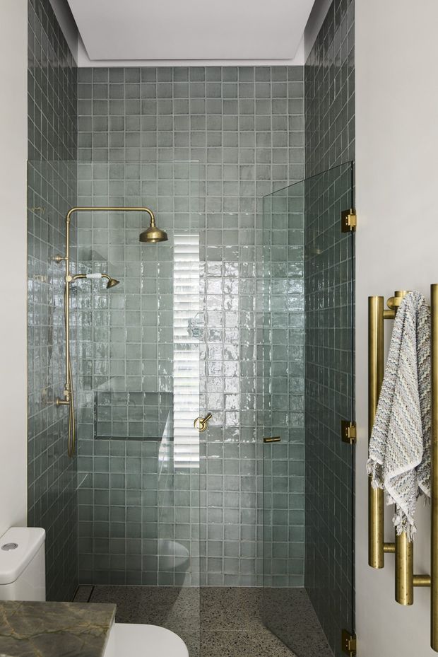 Compact contemporary bathroom featuring a walk-in glass shower enclosed by full-height glossy square sage-grey tiles. Brass/gold shower fittings and towel rails provide warm accents, while a terrazzo-like grey floor and partial white toilet and stone vanity are visible. The shower has a recessed ceiling detail and a minimalist, refined finish.