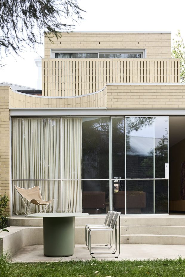 Exterior view of a contemporary brick-clad home with a stepped concrete patio and broad sliding glass doors opening to an interior living area. A curved cream brick privacy wall and vertical screening define the upper terrace, while sparse outdoor furniture sits in the foreground beside a lawn.