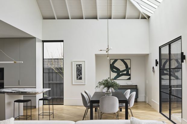 Bright open-plan dining and kitchen interior with a high white vaulted ceiling and exposed rafters. A dark rectangular dining table with mixed upholstered chairs sits centrally, while the kitchen island with a pale stone benchtop is visible at left. Black-framed glazed doors and windows, pale timber flooring, white walls, contemporary artwork, and a built-in display niche create a refined, airy setting.