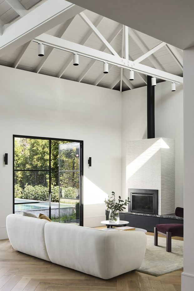 Bright contemporary living room with a dramatic high pitched, exposed white-painted timber truss ceiling. The room features white walls, black-framed glazed doors opening to a garden and pool, a sculptural white curved sofa, pale timber herringbone flooring, a textured white fireplace surround with black flue, and minimalist black cylindrical pendant lights.