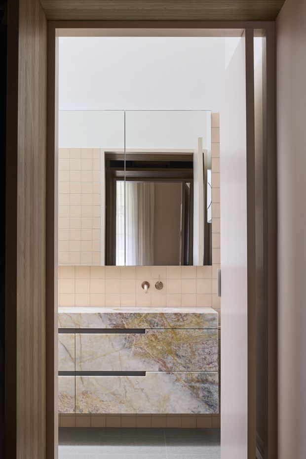 A refined bathroom vanity viewed through a doorway, featuring a floating custom vanity clad in richly veined natural stone, a matching stone benchtop, pale blush-beige square wall tiles, a large mirrored cabinet, and warm timber framing. The compact space has a clean, contemporary feel with brushed warm-metal tapware and soft, natural lighting.