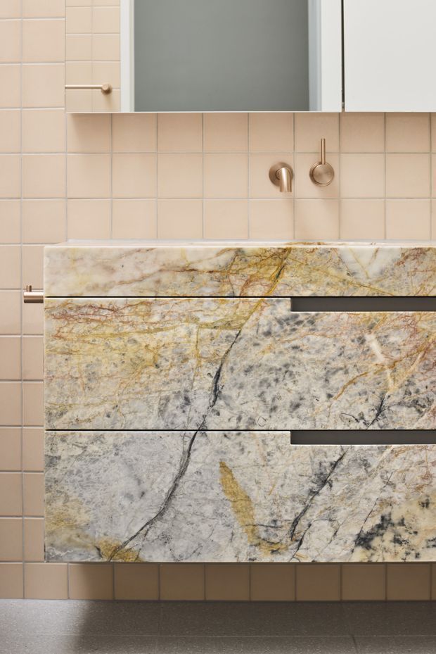 Close-up view of a refined bathroom vanity with dramatic veined natural stone drawer fronts and benchtop, pale beige square wall tiles, an inset mirror, and brushed brass-toned wall-mounted tapware and hardware. The composition highlights the custom joinery and richly patterned stone surfaces.