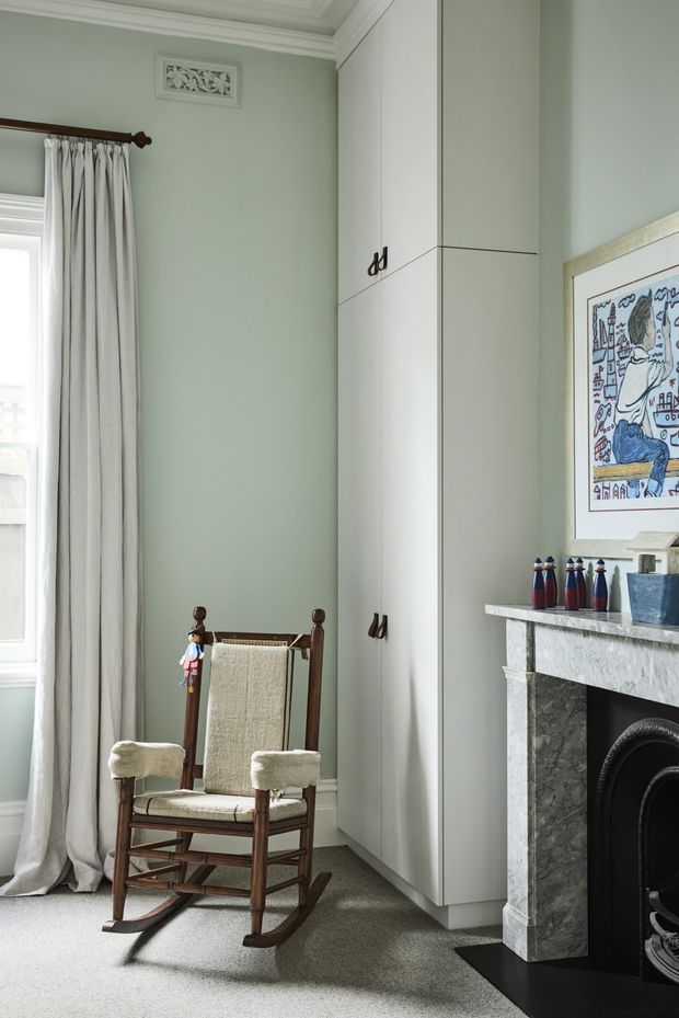 A refined, traditional interior corner featuring a pale sage-green wall, tall white built-in cabinetry, a large window with full-length white curtains, and a wooden rocking chair with woven upholstery. A marble fireplace surround with a dark firebox occupies the right side, topped with small decorative objects and a framed artwork. Ornate ceiling cornices and a decorative wall vent add architectural detail, while grey carpet covers the floor.