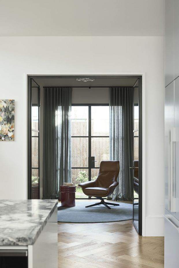 A refined interior view looking through a wide doorway into a small sitting or reading area. The room features a brown leather lounge chair, dark sheer curtains, a black-framed glazed door or window opening to a small garden, and a grey area rug. The foreground has pale herringbone timber flooring and a marble-patterned stone benchtop, with white built-in cabinetry at right. The palette is light and neutral with dark window framing and warm leather accents.