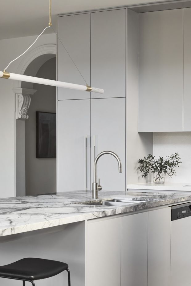 A refined contemporary kitchen with tall light-grey cabinetry, a dramatic veined stone island benchtop incorporating an undermount sink and curved stainless-steel tap, and matching wall cabinetry. A slim linear pendant hangs above the island. An ornate arched architectural opening visible behind the kitchen adds a traditional detail to the otherwise restrained, modern fit-out.