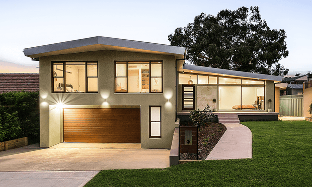 A dusk exterior view of a modern single-storey residence with a prominent flat roof, rendered walls, broad dark-framed windows, and a warm timber-panel garage door. A smaller connected wing extends to the right, with a curved concrete entry path, front lawn, low garden beds, and illuminated facade lighting. The house presents a clean contemporary composition with strong horizontal lines and sheltered outdoor thresholds.