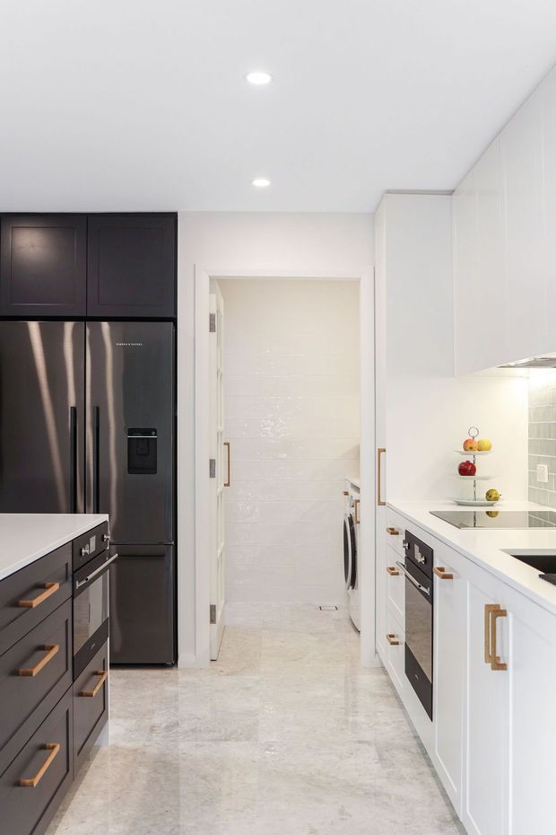 Bright, narrow galley kitchen with a central island, contrasting dark cabinetry and stainless-steel refrigerator on the left, and white shaker-style cabinetry with integrated appliances on the right. A white benchtop, tiled splashback, pale stone-look flooring, brass-toned handles, and recessed ceiling lights create a clean, refined transitional interior. A laundry area is visible through the doorway at the rear.
