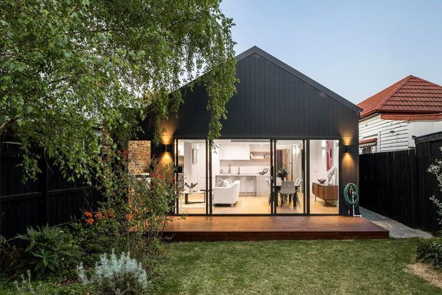 A compact modern backyard pavilion or extension with a steep gabled roof and dark vertical cladding, opening through full-height glazed doors onto a timber deck. The illuminated interior appears to contain a combined living and dining area, while established garden planting and neighboring residential forms frame the exterior.