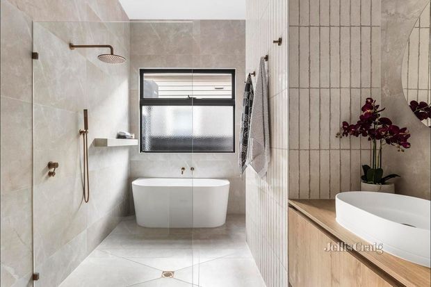 Bright contemporary bathroom featuring a walk-in shower with a rainfall showerhead, freestanding bathtub beneath a frosted window, large pale stone-look wall and floor tiles, and a timber vanity with a second basin visible in the mirror. A vertical tiled feature wall and orchid provide detail.