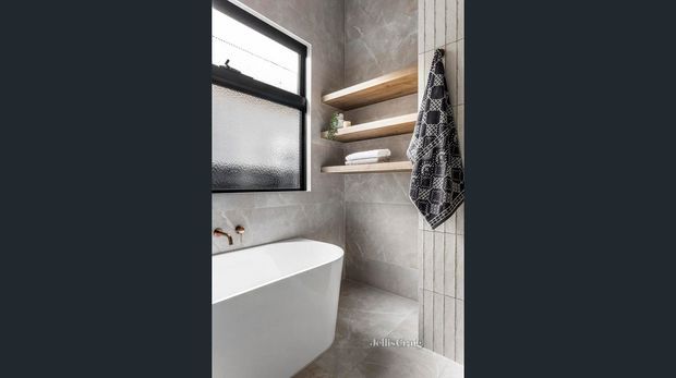 A compact contemporary bathroom featuring a freestanding white bathtub, large-format grey stone-look wall and floor tiles, a black-framed frosted awning window, and open timber shelves with towels and toiletries. A patterned textile hangs beside the shelving. The room has a clean, calm, neutral palette with warm timber accents.