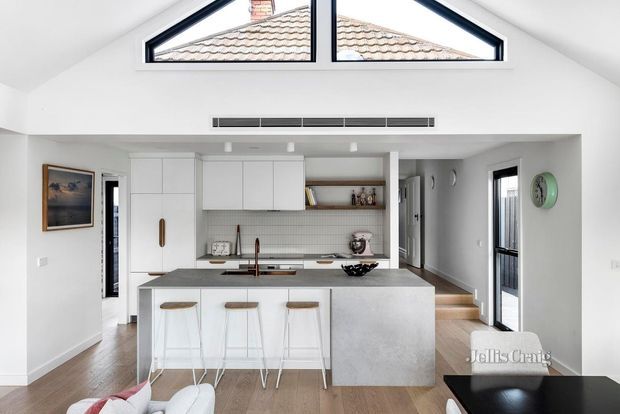 Bright open-plan contemporary kitchen viewed from the living area, featuring a large pale stone island with three stools, white flat-panel cabinetry, a tiled splashback, timber flooring, and a high triangular clerestory window. The space connects to a hallway and adjacent living area, with strong natural light and restrained neutral finishes.