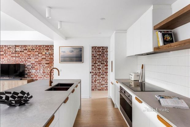 Bright contemporary kitchen with a large pale stone island, white cabinetry, timber shelf accents, white vertical-tile splashback, stainless-steel appliances and warm timber flooring. Exposed red brick walls and a compact living area with television are visible beyond the kitchen, creating a refined modern interior with industrial heritage details.