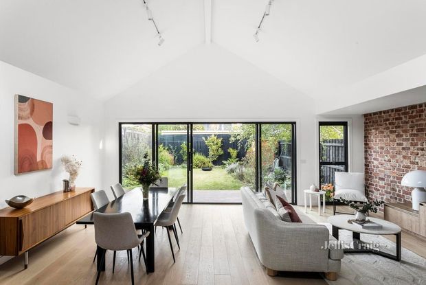 Bright open-plan living and dining room with a pitched white ceiling, pale timber flooring, large black-framed sliding glass doors opening to a garden, exposed red brick feature wall, timber sideboard, dining table and contemporary upholstered seating. Track lighting and restrained furnishings create a clean, airy contemporary interior.