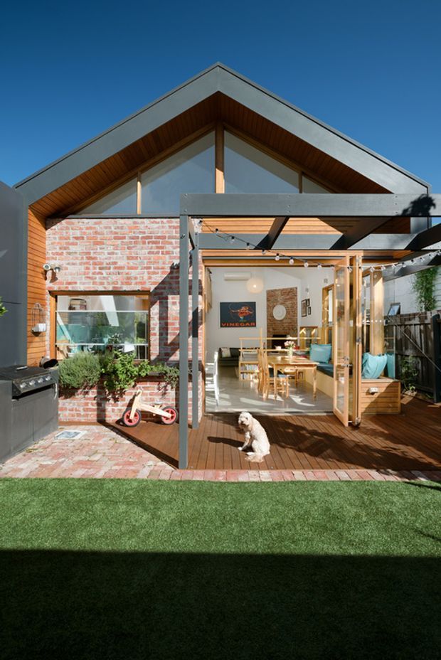 Bright exterior view of a contemporary gable-roofed house opening onto a timber deck and paved courtyard. The facade combines exposed red brick, dark cladding, extensive timber-framed glazing, and a timber-lined gable soffit. A covered pergola with string lights extends over the deck, while the open doors reveal a dining and living area inside. Landscaping, outdoor furniture, a barbecue, and a small dog are visible.