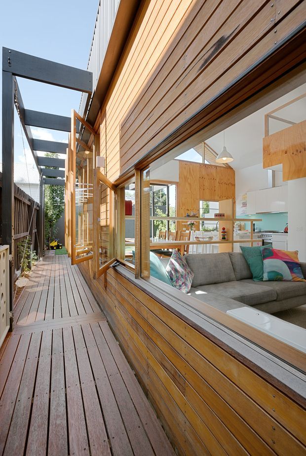 Exterior view along a narrow timber deck and walkway beside a contemporary house. The facade is clad in horizontal natural timber boards with large timber-framed glazing opening onto a bright living room and kitchen visible inside. Dark exposed steel or painted metal overhead beams form a pergola-like structure, while the deck continues toward a small courtyard area. The interior features light timber cabinetry, white surfaces, and a grey sofa visible through the glazing.