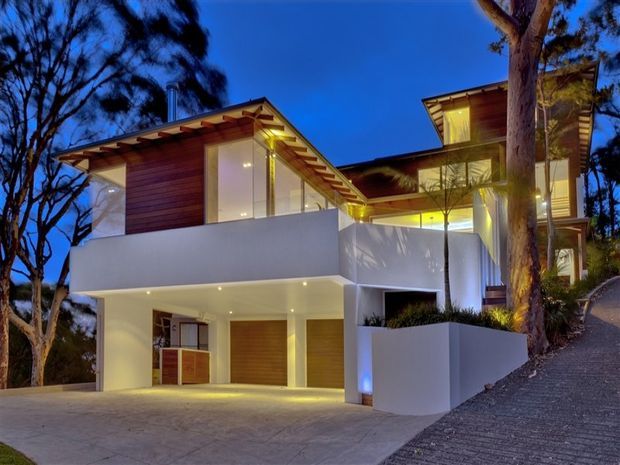 Dusk exterior view of a contemporary multi-level residence set among mature trees on a sloping site. The home features white rendered walls, warm timber-clad upper volumes, broad glazing, exposed roof overhangs, recessed lighting, a large integrated garage, and a concrete driveway with a steep side access path.