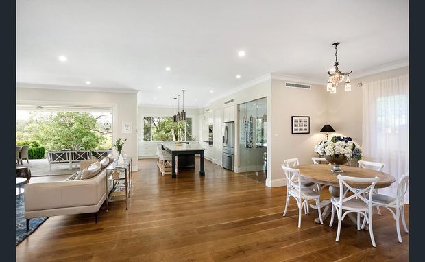 Bright open-plan residential interior combining a living room, dining area and kitchen. The space features warm timber flooring, white walls and cabinetry, large windows, a central kitchen island, a glass-fronted display or storage area, and a traditional dining setting with an ornate pendant light.