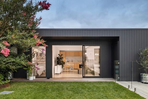A contemporary single-storey home exterior with dark vertical cladding and a flat roof, viewed from a landscaped lawn. Large glazed sliding doors open onto a timber deck, revealing a bright open-plan kitchen and dining area inside. Flowering trees and planting frame the facade, with a glazed balustrade along the deck.