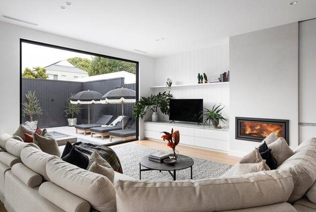 Bright contemporary living room with a large curved cream sectional surrounding a round black coffee table, textured rug, built-in white media cabinetry and shelving, and a recessed fireplace set into a pale feature wall. Full-height glazing opens to a small outdoor seating area with umbrellas and dark fencing. Indoor plants, neutral furnishings, and light timber flooring create a refined, inviting atmosphere.
