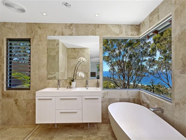 A bright, refined bathroom with large beige natural-stone wall and floor tiles, a wall-mounted white vanity with integrated basin, and a freestanding white bathtub positioned beside expansive corner glazing. The windows overlook trees and a blue coastal or water view. Recessed ceiling lights, a slim ventilation grille, and a louvred window add functional detail.