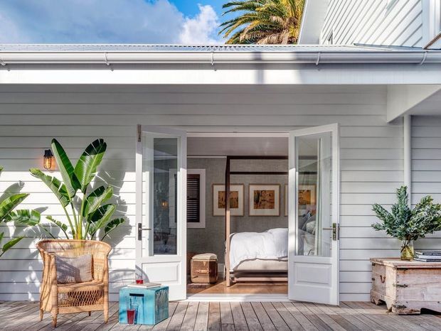 Bright exterior view of a white weatherboard-clad home opening onto a timber deck. Double French doors reveal a bedroom inside, with a bed, framed artwork, and dark timber-framed interior glazing visible. Tropical plants, wicker seating, and a light timber storage chest create a relaxed coastal atmosphere. The image is dominated by the home's exterior facade and deck.