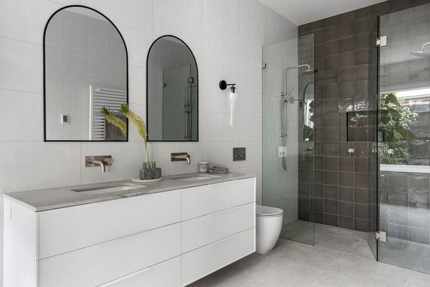Contemporary bathroom with a double white vanity, two arched black-framed mirrors, wall-mounted chrome tapware, and a walk-in glass shower enclosure. The room features large light grey floor and wall tiles, a darker charcoal tiled shower wall, a wall-hung toilet, and a glass door opening toward an exterior garden view.