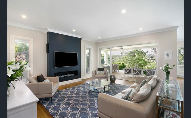 Bright, spacious contemporary living room with cream upholstered sofas and armchairs arranged around a glass coffee table, a large patterned blue rug, warm timber flooring, and a dark feature media wall with wall-mounted television and fireplace insert. Large windows and glazed doors open the room visually to a covered outdoor terrace and leafy surroundings. White walls, recessed lighting, and a glass console contribute to an airy, refined interior.