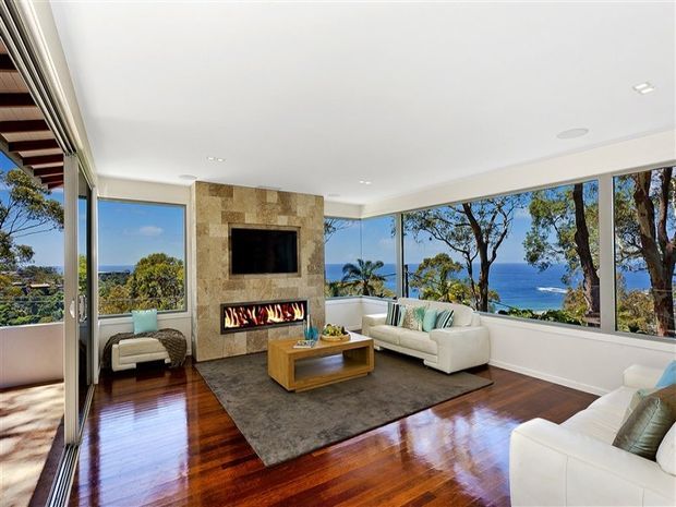 Bright contemporary living room with floor-to-ceiling corner glazing overlooking the ocean and coastal trees. The room features polished dark timber flooring, a stone-clad fireplace wall with an integrated television and linear firebox, white sofas and armchairs, and a large neutral area rug.