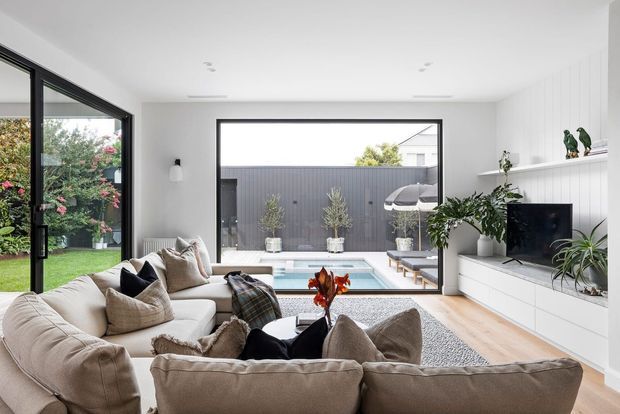 Bright contemporary living room with a large sectional sofa facing a built-in white media wall and television. Full-height black-framed glazing opens the room visually to a compact courtyard and swimming pool, while light timber flooring, white vertical wall panelling, neutral furnishings, and indoor plants create an airy, refined interior.