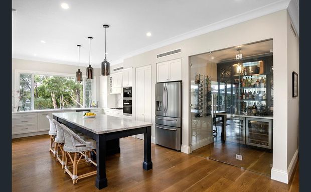 Bright, spacious contemporary kitchen with a large marble-look island, white shaker-style cabinetry, stainless-steel appliances, pendant lighting, and warm timber flooring. A glazed walk-in wine room or bar occupies the right side, with extensive bottle storage, display shelving, and a wine fridge. The kitchen opens toward a leafy outdoor view.
